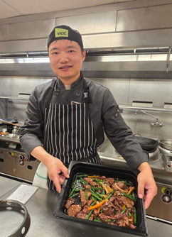 Chef in black uniform holding a tray of stir-fried beef with green beans in a professional kitchen.