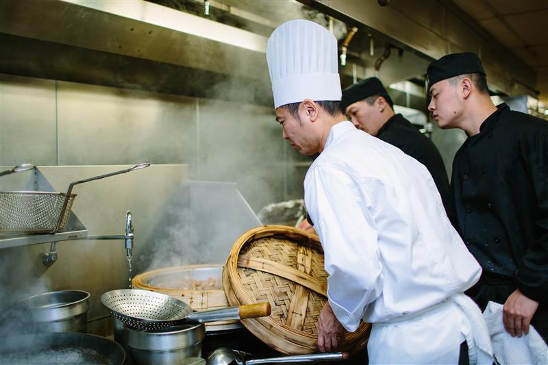 Professional chefs working in a commercial kitchen with steam rising from pots and a large bamboo steamer basket being handled.