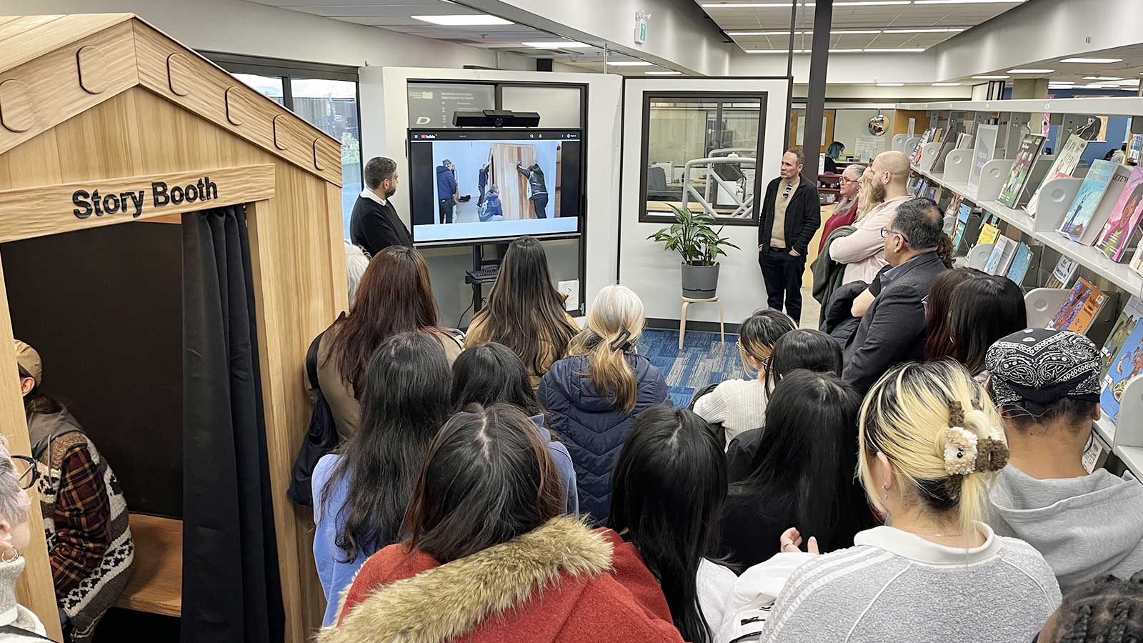 A crowd of people gathers in a library for the demonstration of an interactive wooden "Story Booth" installation.