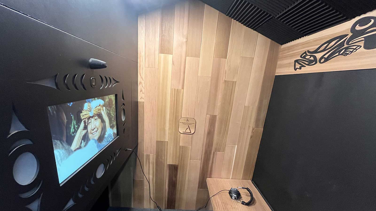 The interior of a wood-paneled "Story Booth" features a digital screen displaying a video and traditional Indigenous-style carvings integrated into the black acoustic walls.